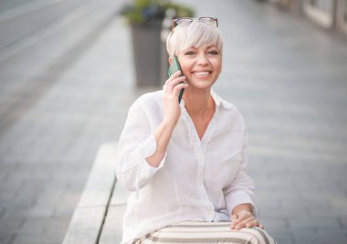 Cep telefonuyla konuşan, kentteki bankta dinlenen orta yaşlı güzel bir kadın. Yetişkin kadın akıllı telefondan konuşuyor. Dışarıda, dışarıda.