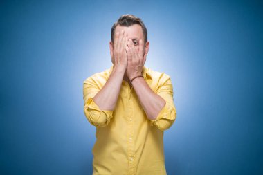 Zirve. Yüzü kararmış. Yarı uzunlukta genç bir adam elleriyle yüzünü kapatıyor. Mavi arka planda utanç, sarı gömlekli. Utangaç adam. Stüdyo çekimi