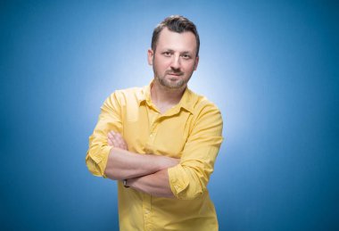 Mavi arka planda çapraz kollu, sarı tişörtlü, pozitif genç bir adamın portresi. Kameraya bakan neşeli adam. Stüdyo çekimi