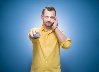 Sıkıcı genç adam televizyon izliyor ve elinde uzaktan kumandayla mavi arka planda, sarı gömlekli. Sıkıcı adam kameraya bakıyor. Stüdyo çekimi