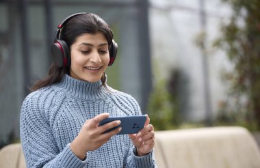 Açık havada kablosuz kulaklık takan genç bir kadın cep telefonuyla film ya da film izliyor.