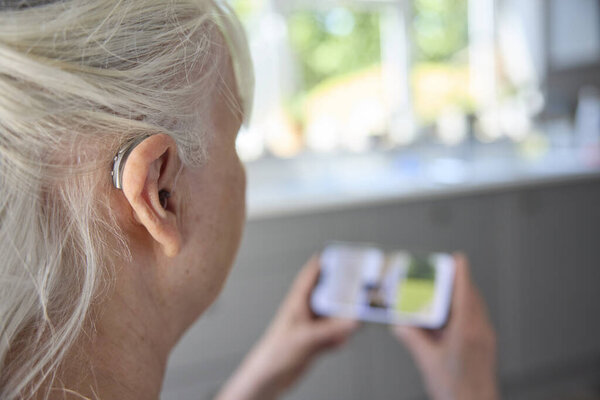 Крупный план пожилой женщины, носящей за ухом слуховой аппарат или всплеск помощи с мобильного телефона дома