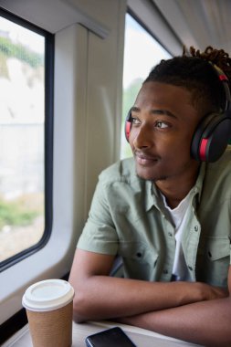 İşe gidip gelen genç adam, trende oturuyor, kablosuz kulaklık takıyor ve camdan dışarı bakıyor.