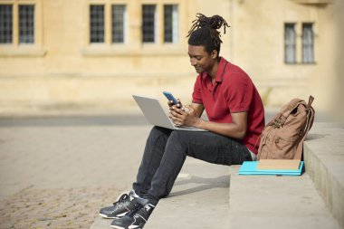 Oxford 'daki üniversite binasının dışındaki basamaklarda oturan erkek öğrenci laptopta mesajları veya cep telefonundaki sosyal medyayı kontrol ediyor.