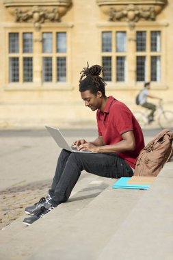Oxford 'daki üniversite binasının dışındaki basamaklarda laptopta çalışan erkek öğrenci.