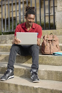 Oxford 'daki üniversite binasının dışındaki basamaklarda oturan erkek öğrenci laptopta mesajları veya cep telefonundaki sosyal medyayı kontrol ediyor.