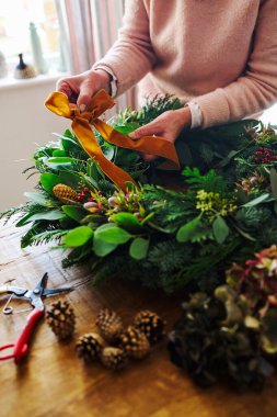 Close Up Of Woman Making And Decorating Festive Christmas Wreath On Table At Home Fastening Bow Made From Ribbon clipart