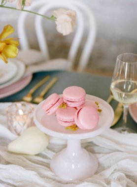 Pembe badem kurabiyeleri beyaz bir kaideye güzelce yerleştirilmiş çiçeklerle süslenmiş güzel bir yemek masası ve zarif bir cam eşyayla çevrili. Bahar kutlaması için mükemmel..