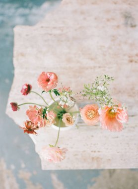 Pembe ve beyaz tonlarında narin çiçekler açık renkli bir masadaki bir vazoda ustalıkla düzenlenir. Doğal ışık, her çiçeğin karmaşık yapraklarını vurgular..