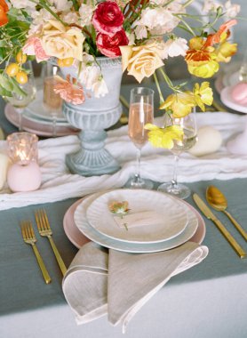 Güzelce düzenlenmiş bir yemek masasında zarif sofra takımı ve ışıl ışıl gözlüklerle çevrili çarpıcı bir çiçek süsü bulunur. Yumuşak renkler neşeli bir ambiyansa katkıda bulunur..
