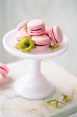 Hassas pembe badem kurabiyeleri, yeşil çiçeklerle süslenmiş beyaz kaideye dizilir. Tatlıların cazibesini ve zarafetini arttırmak için ayarlar yumuşak bir şekilde aydınlatılır..