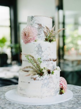 Güzelce hazırlanmış üç katlı bir pasta, doğal ışık ve yeşillikle dolu şık, modern bir mekanda sergilenen taze çiçekler ve yeşillikle süslenmiş pürüzsüz bir beyaz kaplama sergiliyor..