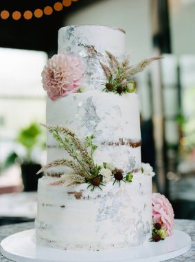 Çok güzel tasarlanmış üç katlı bir düğün pastası dairesel bir zeminde duruyor, taze çiçekler ve yeşilliklerle süslenmiş, şık bir resepsiyon salonunda. Yumuşak doğal ışık sahneyi güçlendirir..