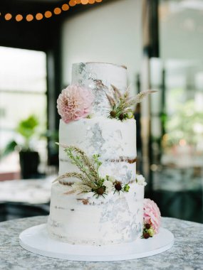 Büyüleyici üç katlı düğün pastası zarif beyaz süslemeler, narin çiçekler ve yeşillikler içerir. Hepsi de hoş, romantik bir ortamda, yumuşak ışıklandırmayla sergilenir..