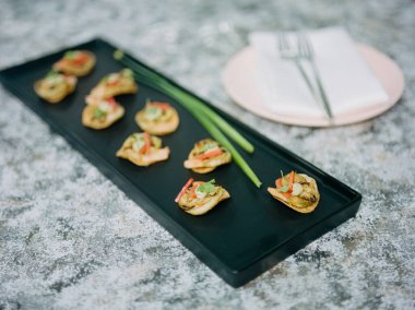 Parlak siyah bir tabakta her birinin üzerinde taze malzemeler bulunan çeşitli lezzetli mezeler sergilenir. Yanında çatal bıçak olan pembe bir tabak, lüks bir yemek deneyimi olduğunu gösteriyor..