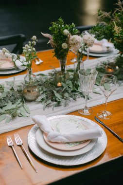 Güzelce düzenlenmiş bir yemek masası yumuşak pembe tabaklar, kristal cam eşyalar ve çiçek süslemeleri ile yemyeşil bir ortam oluşturur..