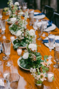 Uzun tahta bir masa beyaz çiçekler, yeşillik ve mumlardan oluşan zarif bir düzenle süslenir. Parlak ve davetkar bir ortam. Tabaklar ve kadehler zarifçe yerleştirilmiş..