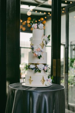 Güzel tasarlanmış dört katlı bir düğün pastası sıcak ışıklarla dolu şık bir mekanda zarif bir şekilde taze çiçekler ve altın aksanla süslenmiş bir masada sergileniyor..