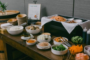 A vibrant poke bar displays an array of fresh ingredients including rice, seafood, and vegetables. Guests are invited to create personalized bowls at this casual dining setup during a gathering. clipart