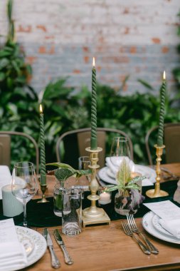 Uzun yeşil mumlar, cam eşyalar ve dekoratif bitkilerle güzel düzenlenmiş bir yemek masası modern bir restoran ortamında misafirperver bir ortam yaratıyor..
