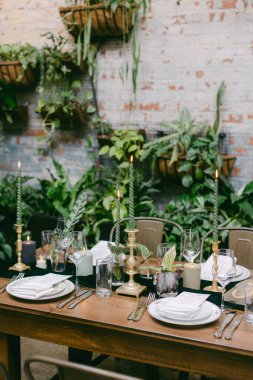 Güzel düzenlenmiş bir yemek masası yeşil mumlar, cam eşyalar ve canlı bitkilerle çevrili tabaklar içerir. Kurulum öğleden sonra için sıcak bir atmosfer yaratıyor..