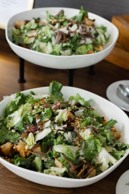 Çağdaş bir yemek salonunda, ahşap bir masada karışık yeşillikler, çıtır çıtır malzemeler ve canlı renklerle dolu enfes bir kase taze salata sergileniyor..