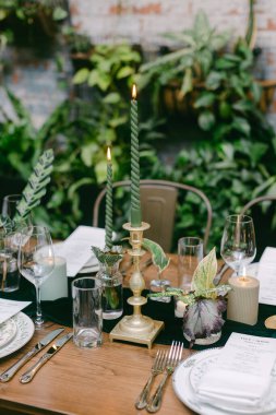 Çok güzel düzenlenmiş bir yemek masasında altın bir şamdanın üzerinde yeşil mumlar, bitkiler ve zarif camlarla çevrili, sıcak ve davetkar bir yemek deneyimi yaratıyor..