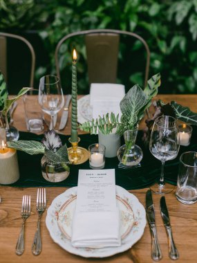 Güzel düzenlenmiş bir yemek masası ayrıntılı yer ayarları, taze yeşillik, yumuşak mum ışığı, ve hassas cam eşyalar, akşam yemeği için sıcak bir ortam yaratır..