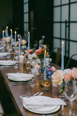 Pastel çiçeklerin, mavi mumların ve zarif sofra takımlarının olduğu güzel bir yemek masası. Dekor, şık ve çağdaş bir mekanda özel bir kutlama için tasarlandı..