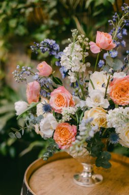 Göz kamaştırıcı pastel güller, beyaz çiçekler ve yaban çiçekleri metalik bir vazoda zarif bir şekilde dizilmiş ahşap bir yüzey üzerinde duruyor, canlı yeşilliklerle çevrili, taze bir ambiyansı çağrıştırıyor..