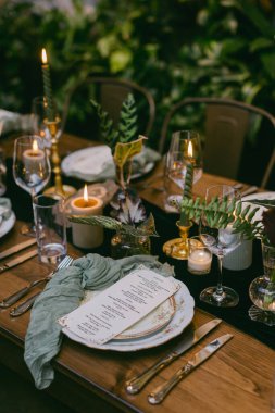 Güzel düzenlenmiş bir yemek masasında zarif tabaklar, bardaklar ve mumlar bulunur. Yeşilliklerle çevrili. Özel bir toplantı için rahat bir ortam yaratırlar..