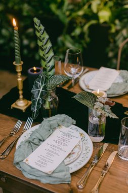 Güzel düzenlenmiş bir yemek masasında zarif tabaklar, yumuşak mum ışığı ve yemyeşil bir ortam var. Konuklarla unutulmaz bir yemek deneyimi için ortam hazırlıyorlar..