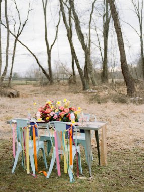 Çok güzel düzenlenmiş bir açık masa canlı çiçekler ve sandalyelere takılmış renkli kurdeleler sergiliyor. Bu düzen, misafirleri doğadaki bahar toplantısının tadını çıkarmaya davet ediyor..