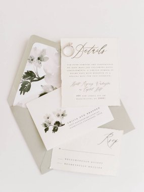 Çok güzel düzenlenmiş bir düğün davetiyesi süitinde yumuşak tonlarda narin çiçekli sanat eserleri, açık renkli bir yüzey üzerinde RSVP ve ayrıntılı kartlar sergileniyor..