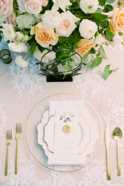 Güzel düzenlenmiş bir yemek masasında altın çatal bıçak ve dekoratif menüsü olan zarif bir tabak bulunur. Taze güller ve yeşillik göz kamaştırıcı bir ortam oluşturur..