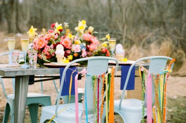 Güzel dekore edilmiş açık hava masasında güller ve nergislerle süslenmiş parlak bir çiçek sergileniyor. Sandalyelerden sarkan renkli kurdeleler, toplantı için şenlikli bir atmosfer yaratıyor..