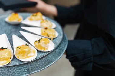 Bir sunucu, beyaz kaşıklarda zarif bir şekilde yerleştirilmiş enfes aperatiflerle dolu bir tepsi sunar. Mekan, misafirlerin yemek ziyafetlerinin keyfini çıkardıkları sofistike bir olaya işaret ediyor..