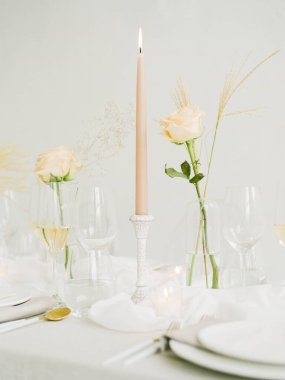 Güzelce düzenlenmiş bir yemek masasında zarif güller ve camlarla çevrili uzun bir mum bulunur. Yumuşak ışıklandırma özel bir durum için sıcak ve davetkar bir ortam yaratır..