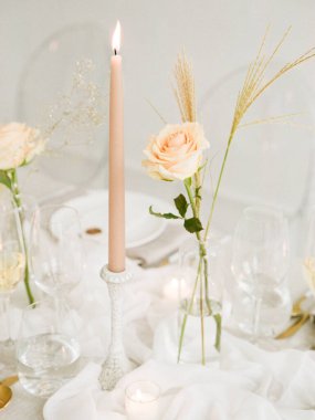 Yumuşak bir gül, altın buğday ve yanan bir mum içeren hassas masa düzenlemesi samimi bir akşam yemeği partisi için sıcak bir ortam yaratır. Gözlükler ve tabaklar zarif stili tamamlar.