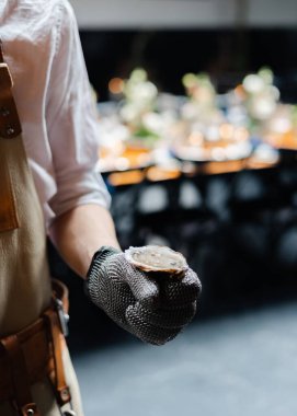 A chef in a white shirt and gloves holds a freshly shucked oyster, ready to serve at a fine dining establishment. clipart