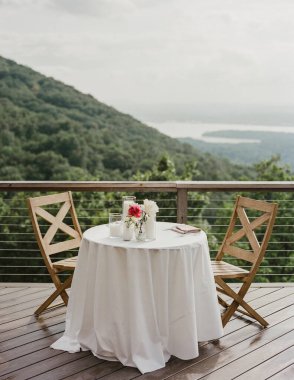 İki kişilik küçük yuvarlak bir masa, taze çiçeklerle, bulutlu bir gökyüzünün altında yemyeşil bir dağ manzarasına bakıyor..