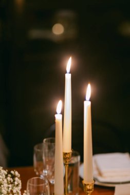 Üç uzun, beyaz mum, loş, samimi bir ortamda cam eşyayla rahat bir yemek masasını aydınlatır..