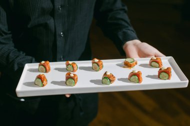 Bir garson bir tepsi renkli suşi sunar içinde taze salatalık ve çıtır çıtır soslar vardır..