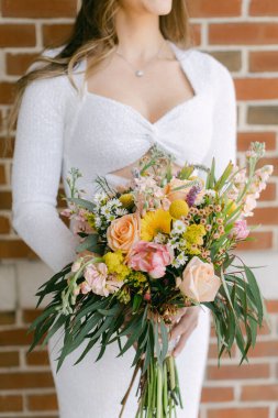Beyaz elbiseli gelin, tuğla bir arka planda poz verirken renkli bir çiçek buketi sergiliyor.