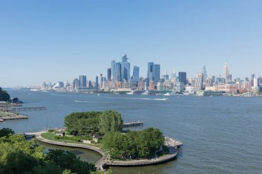 Sakin bir nehir canlı bir New York şehrinin üstünden akar, yüksek binalar ve kıyı boyunca yemyeşil alanlar sergilenir..