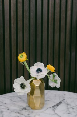 Beyaz şakayık çiçekleri ve sarı çiçekleri içeren canlı bir koleksiyon modern altın bir vazoda koyu ahşap panellerle çevrili mermer bir masada duruyor..