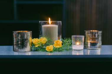 Yumuşak mum ışığı çeşitli tutuculardan titreşiyor, canlı sarı çiçekler ve yeşil çiçeklerle çevrili basit bir masanın üzerinde, odanın sıcak, davetkar atmosferini arttırıyor..