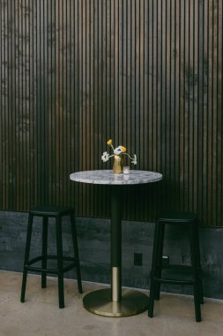 Küçük yuvarlak bir masa, şık ahşap bir duvara dayanmış vazo dolusu taze çiçeklerle süslenir. Masanın etrafında iki siyah tabure, hoş bir atmosfer yaratıyor..