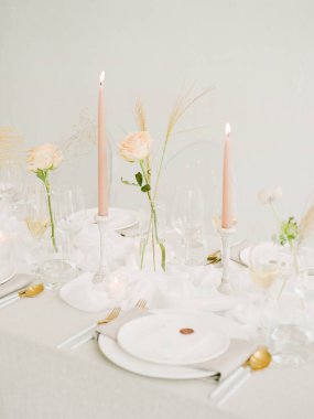 Güzel düzenlenmiş bir yemek masasında soluk pembe mumlar ve narin çiçek sapları bulunur. Zarif sofra takımı yumuşak, sıcak aydınlatma altında parlıyor ve sakin bir atmosfer yaratıyor..