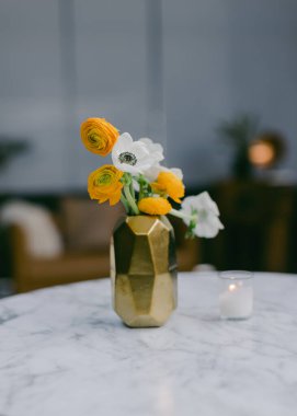 Sarı ve beyaz çiçeklerden oluşan canlı bir buket zarif bir altın vazoda duruyor, arka plandaki yumuşak dekorla aydınlatılmış mermer bir masanın üzerinde..